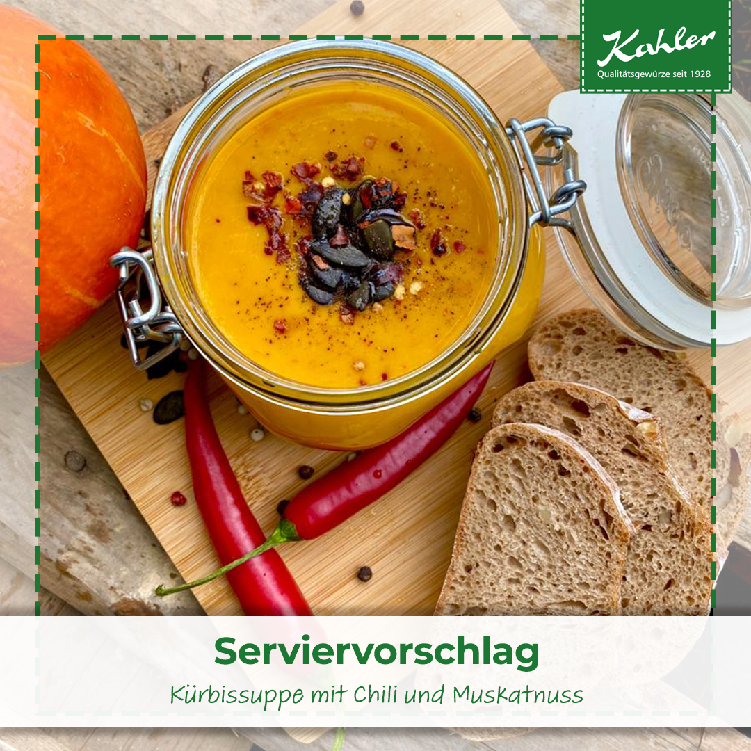 Glas mit Kuerbissuppe, garniert mit Kuerbiskernen und Gewuerzen, auf Holzbrett mit zwei roten Chilischoten und drei Scheiben Vollkornbrot.