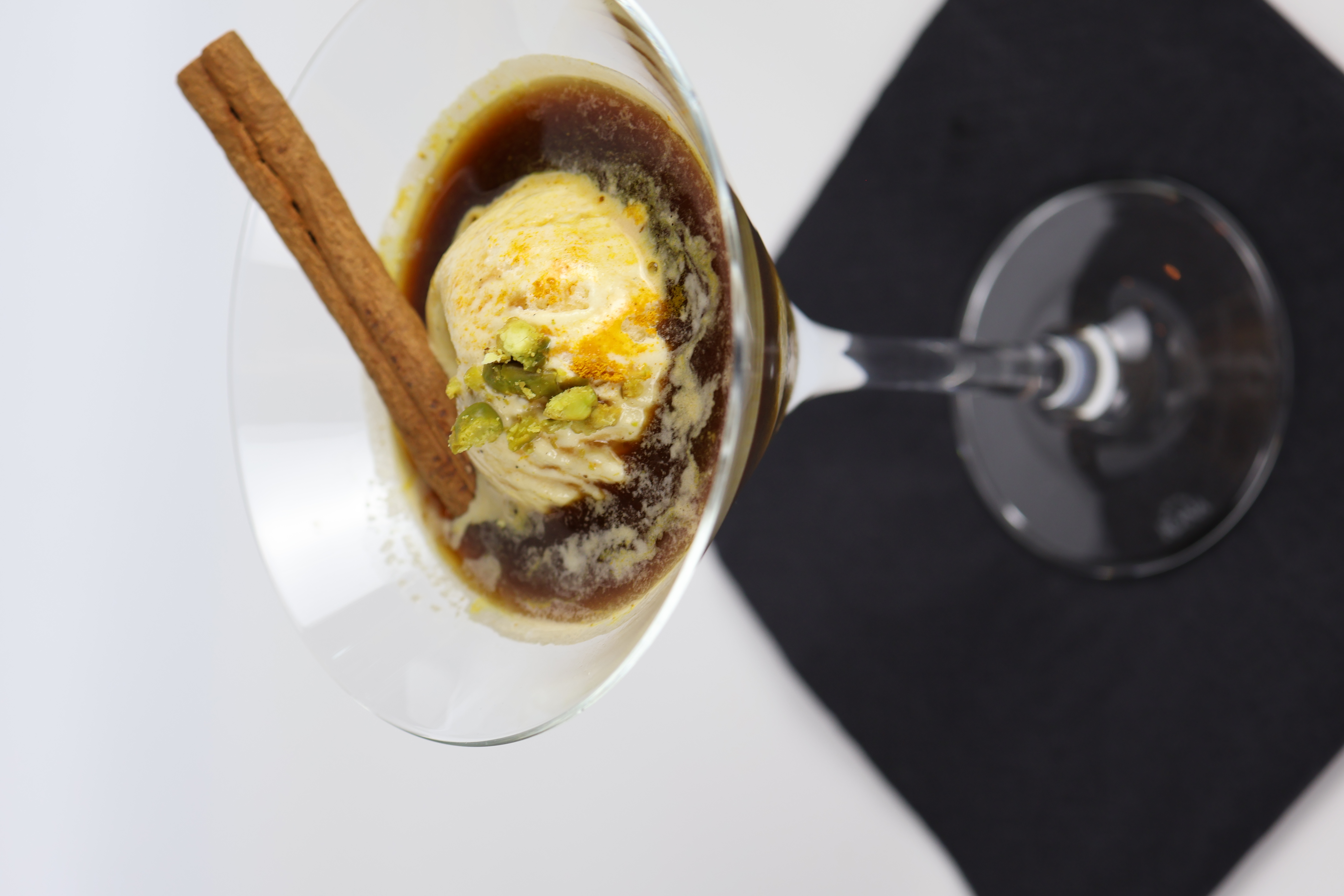 Cocktailglas mit einer Kugel Vanilleeis in dunkler Flüssigkeit, garniert mit einer Zimtstange und gehackten Pistazien