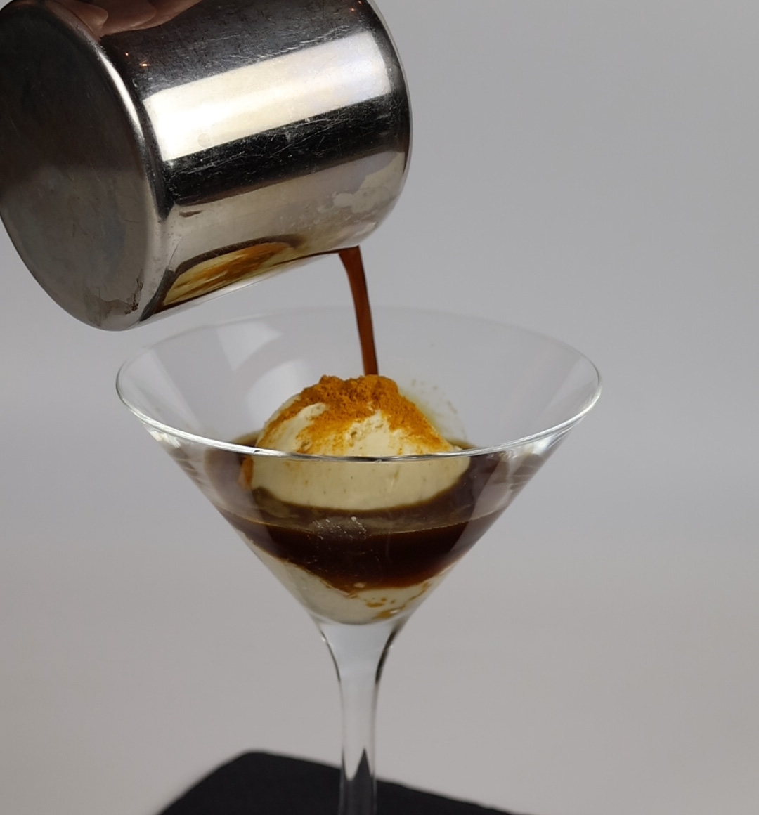 Cocktailglas mit einer Kugel Vanilleeis, das mit einer braunen Flüssigkeit aus einem Metallbehälter übergossen wird